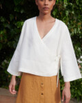Model wearing a loose-fitting white linen top with a deep V-neck and long kimono sleeves paired with a camel linen skirt with metal snap buttons at the front