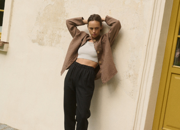Model leaning against a wall wearing high waisted black heavy linen pants and brown linen jacket with wooden buttons