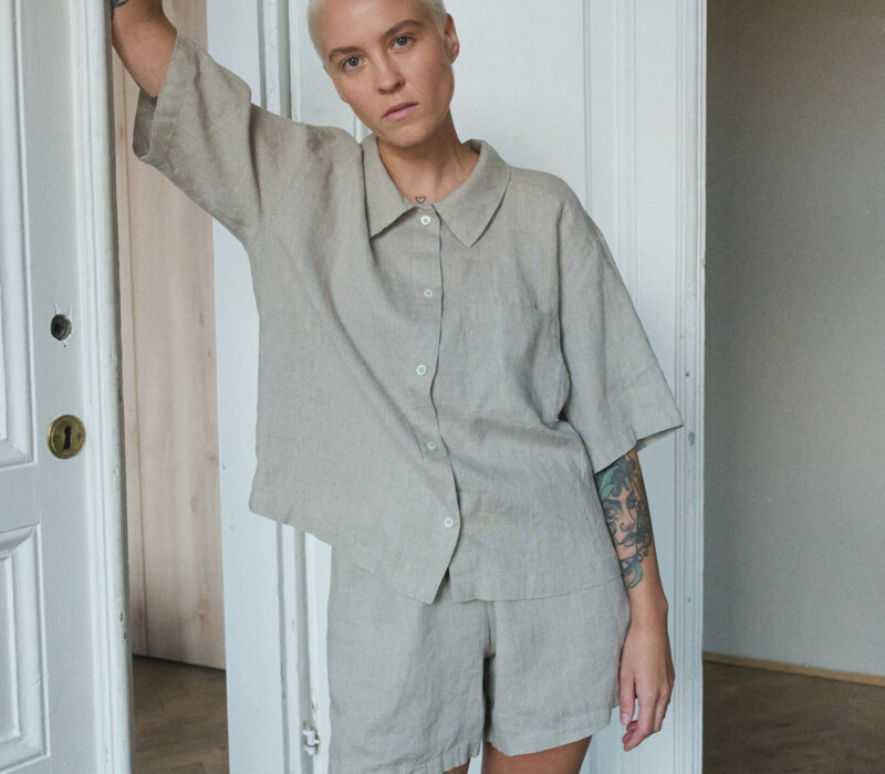 Model wearing a relaxed, natural gray linen two-piece pajama set, including a loose-fitting, short-sleeve button-up shirt with a wide collar and a straight hem that falls just above the waist, and matching shorts in a simple, casual design, reaching mid-thigh