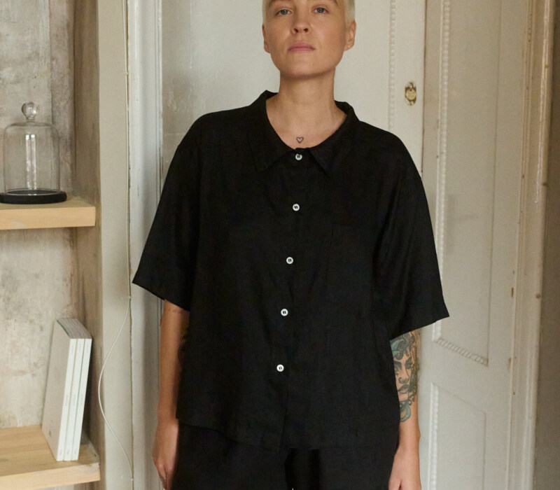 Woman wearing a relaxed black linen two-piece set, featuring a loose short-sleeve button-up shirt and mid-thigh shorts, offering a minimalist and casual look