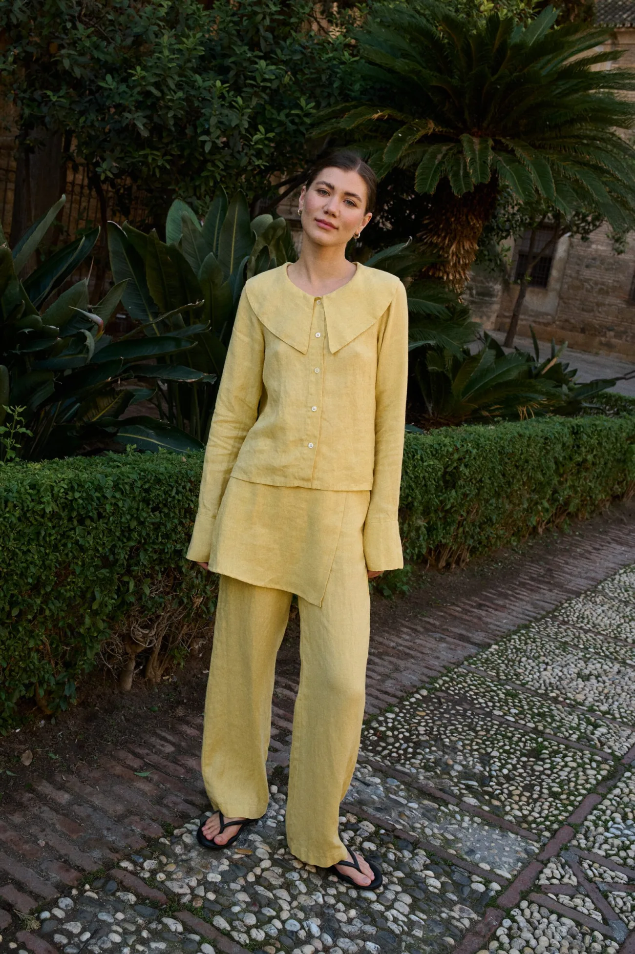 Model in a butter-yellow linen three-piece outfit featuring a tailored button-front shirt with distinctive oversized collar, asymmetrical layered skirt panel, and relaxed wide-leg trousers
