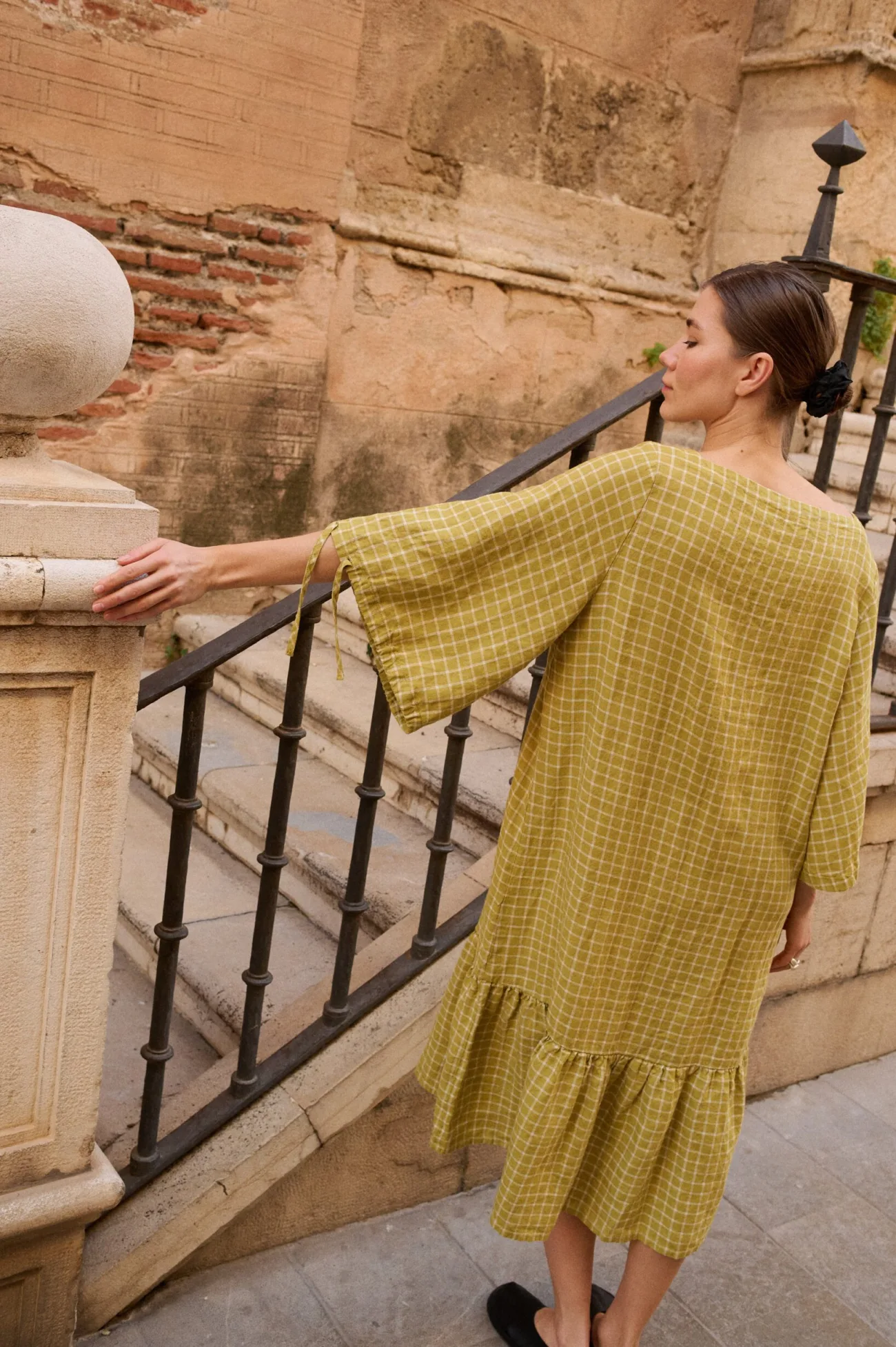 Model seen from the back, wearing a pickle checks linen dress with wide, flared three-quarter length sleeves that feature tie details and a dropped waistline with a voluminous ruffle hem