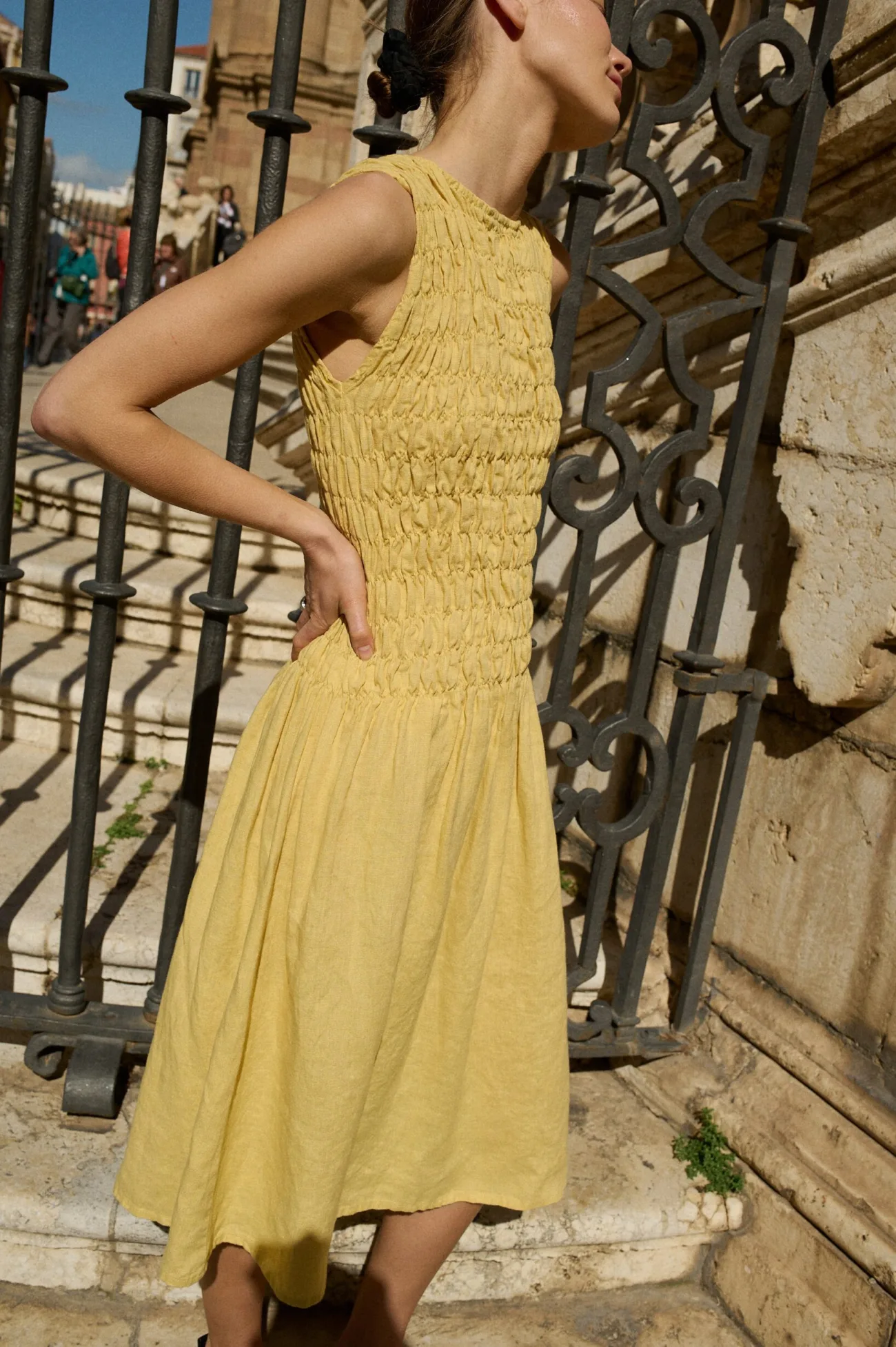 Model wearing a sleeveless butter-yellow linen dress with a smocked bodice that gracefully flows into a midi-length skirt, creating an elegant and airy silhouette