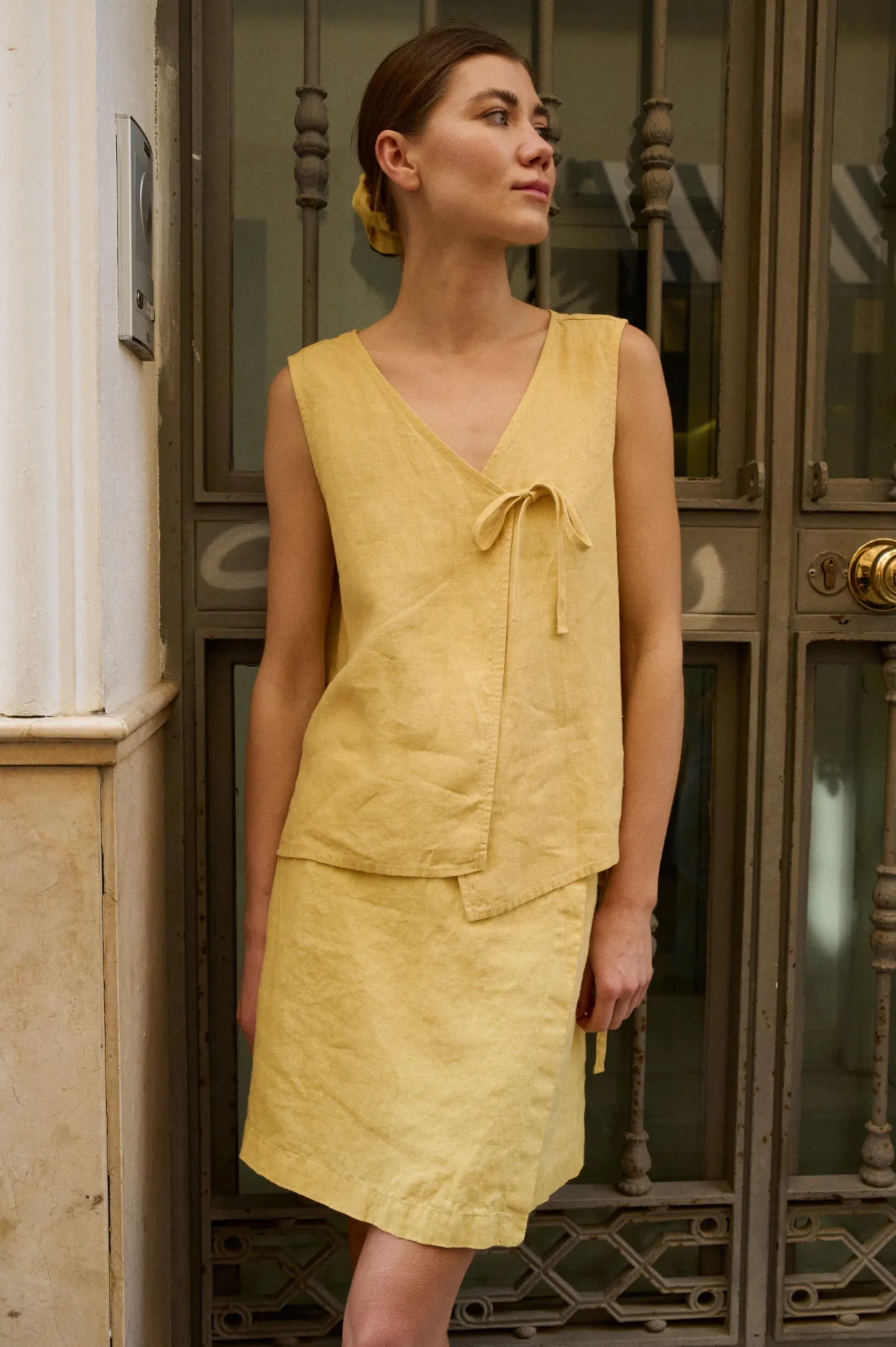 Model standing in front of an ornate glass doorway, wearing a butter-colored linen top with an asymmetrical wrap-style and tie detail at the side, paired with a matching skirt, creating a layered effect