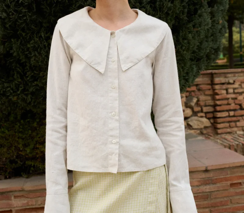 Model wearing a white cotton blend blouse with an oversized Peter Pan collar, button-down front, and full-length sleeves with subtle cuffs, paired with a pale apple checks linen wrap skirt featuring side tie closures and a subtle A-line shape