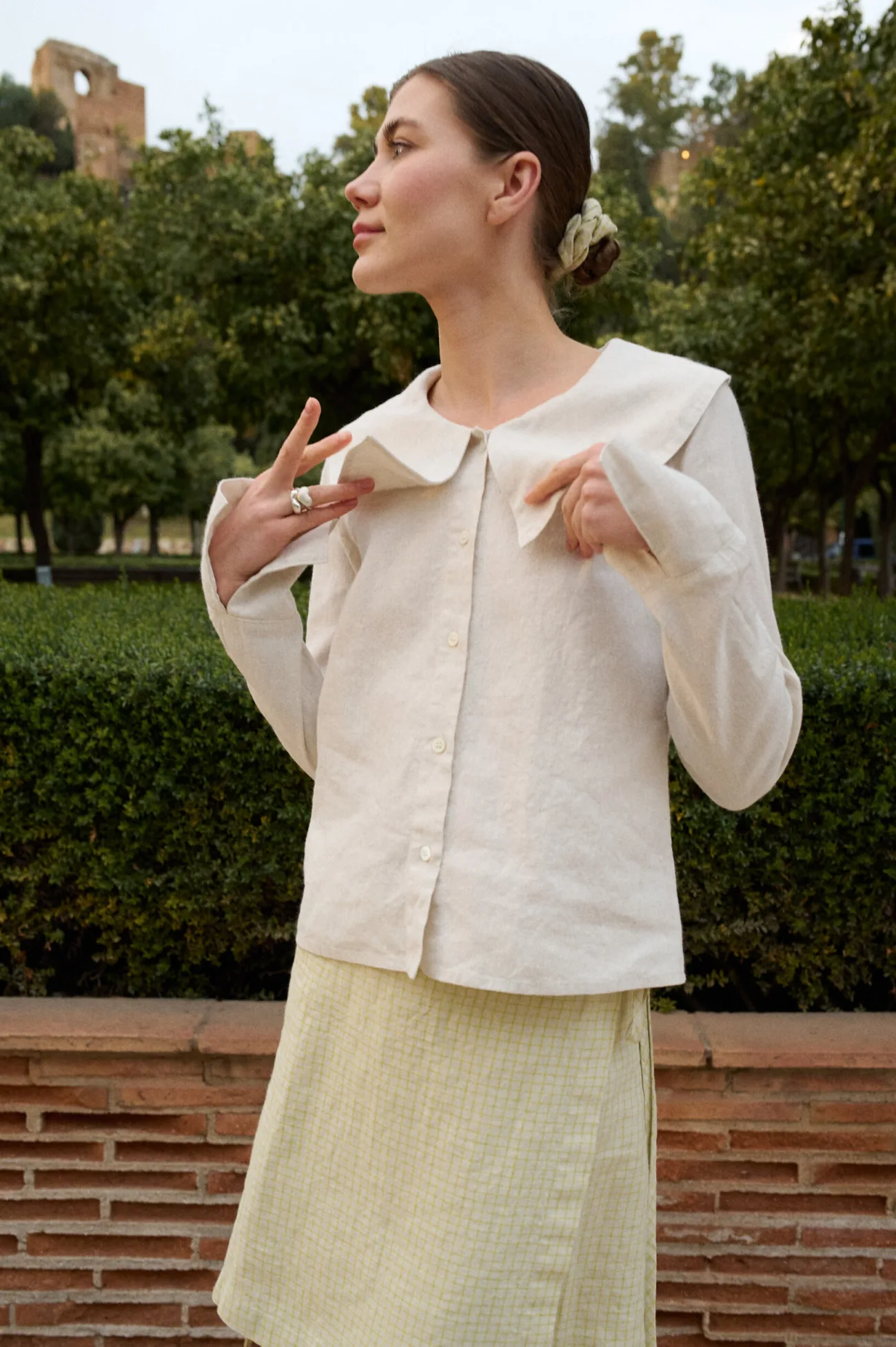 Model wearing a white cotton-blend blouse with an oversized Peter Pan collar, button-down front, and full-length sleeves with slits, paired with a pale apple checks linen wrap skirt
