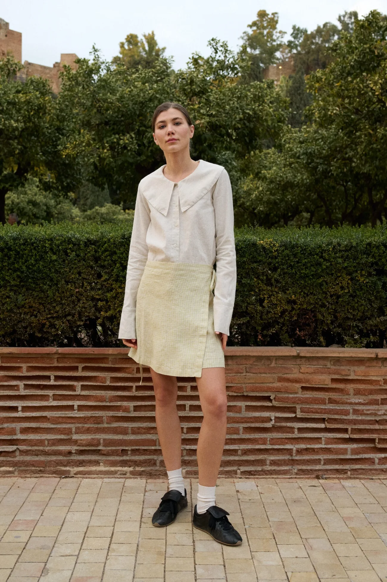 Model wearing a light yellow checkered linen wrap skirt with side ties, paired with a natural grey button-up top featuring an oversized collar
