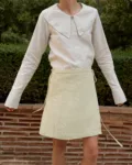 Model in a garden setting, wearing a natural grey button-up top with an oversized collar, tucked into a light yellow checkered linen wrap skirt with side ties