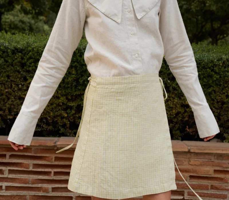 Model in a garden setting, wearing a natural grey button-up top with an oversized collar, tucked into a light yellow checkered linen wrap skirt with side ties