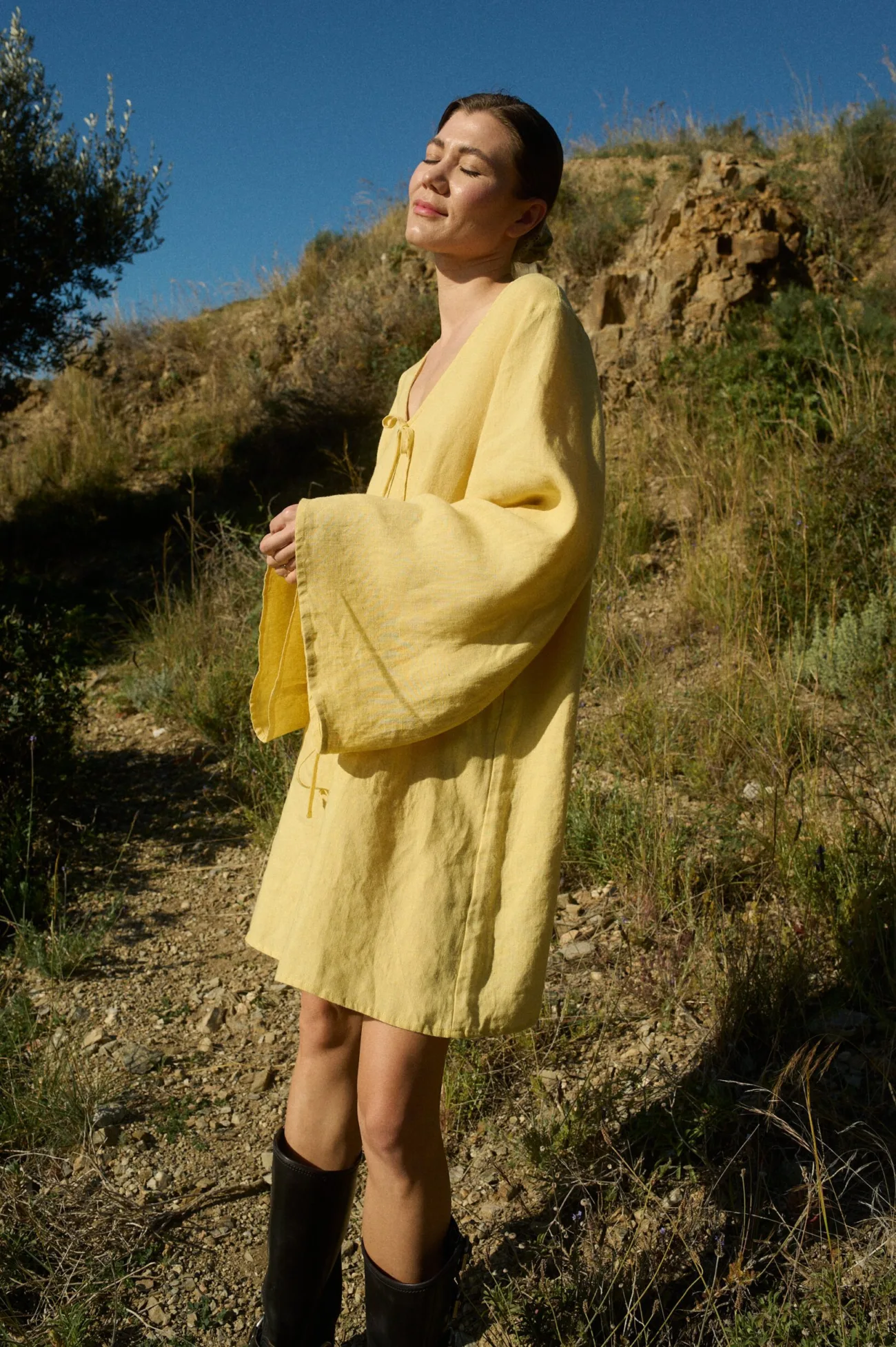 Model wearing a butter-colored mini linen dress featuring a deep V-neckline, oversized A-line silhouette, flowy wide sleeves, and four decorative front tie-bows