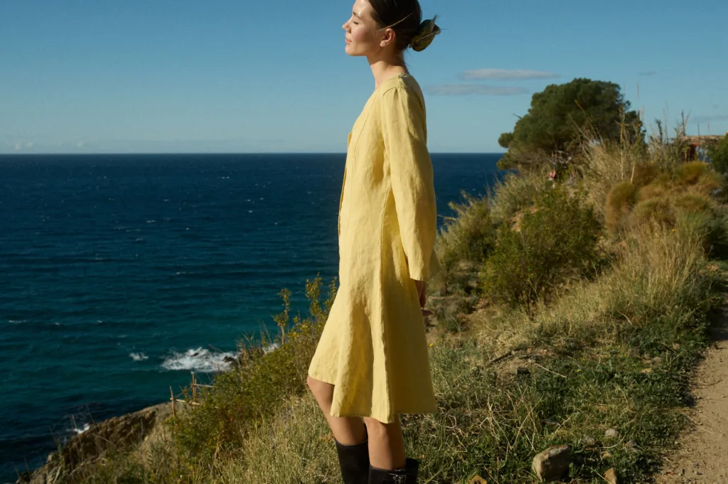 Model pictured from the side in a butter-colored linen dress featuring vertical linen panels and godet inserts that create a flowing, A-line silhouette