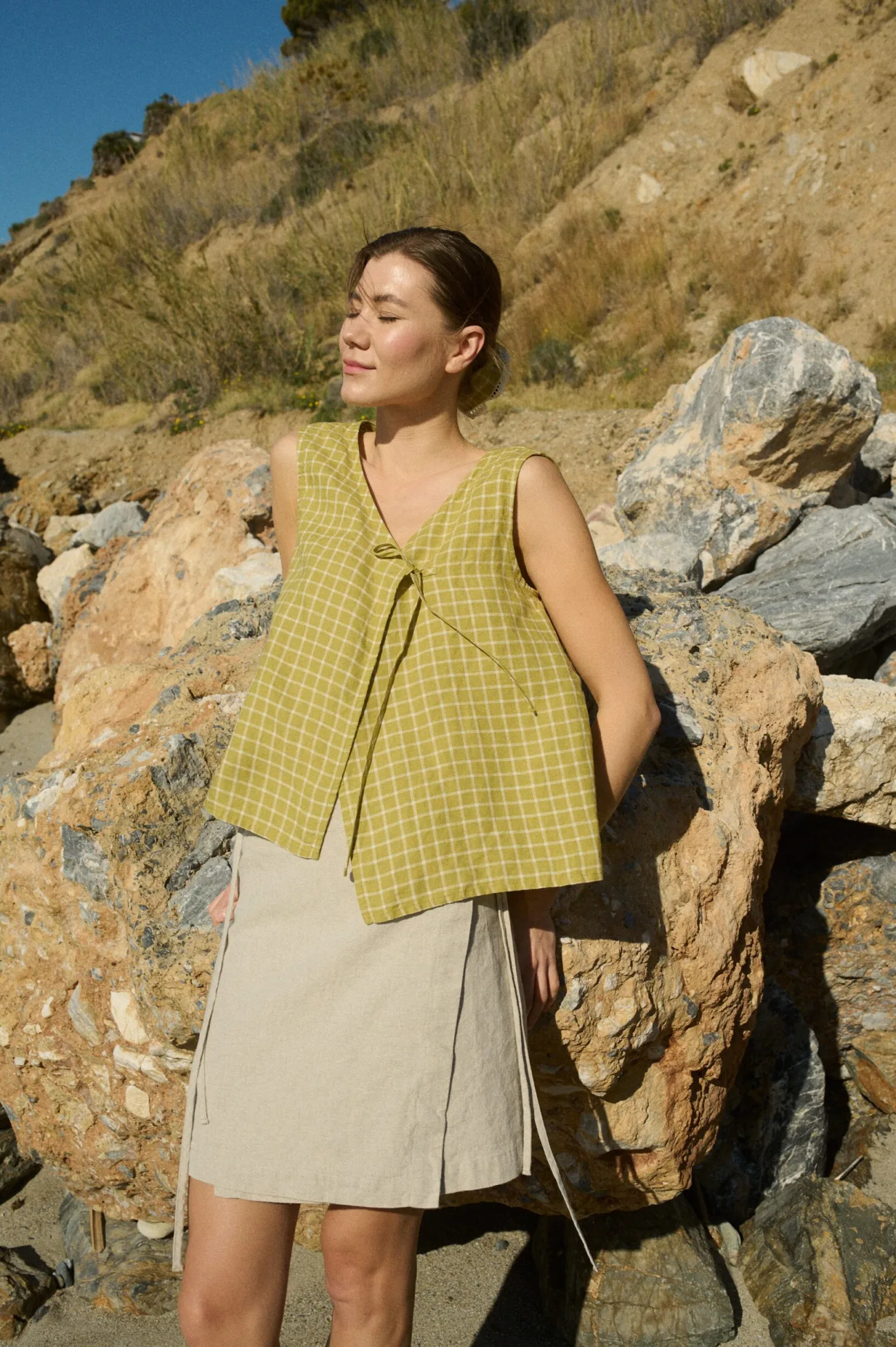 Model in a sleeveless pickle checks linen top with a V-neck and relaxed fit, complemented by a light beige natural cotton blend wrap skirt, fastened at the sides and falling to a mini length