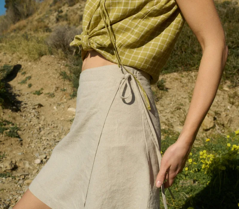 Model dressed in a sleeveless pickle checks linen top with a front tie, paired with a natural cotton blend wrap skirt that falls above the knee