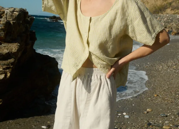 Model wearing a short-sleeved, button-up top in apple checks linen with a scoop neckline, slightly relaxed fit, and full-length buttons down the center front, paired with off-white wide-leg cotton pants with an elastic waistband