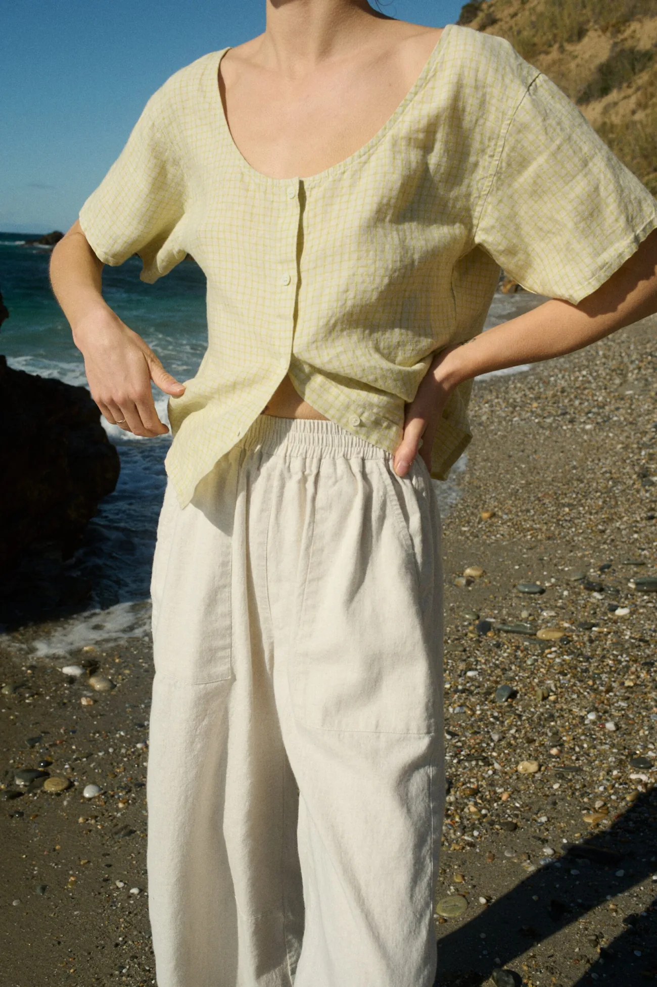 A pale yellow checkered short-sleeve linen button-up with a scooped neckline paired with cream linen cotton blend wide-leg pants featuring an elastic waistband and side pockets, worn at a seaside setting