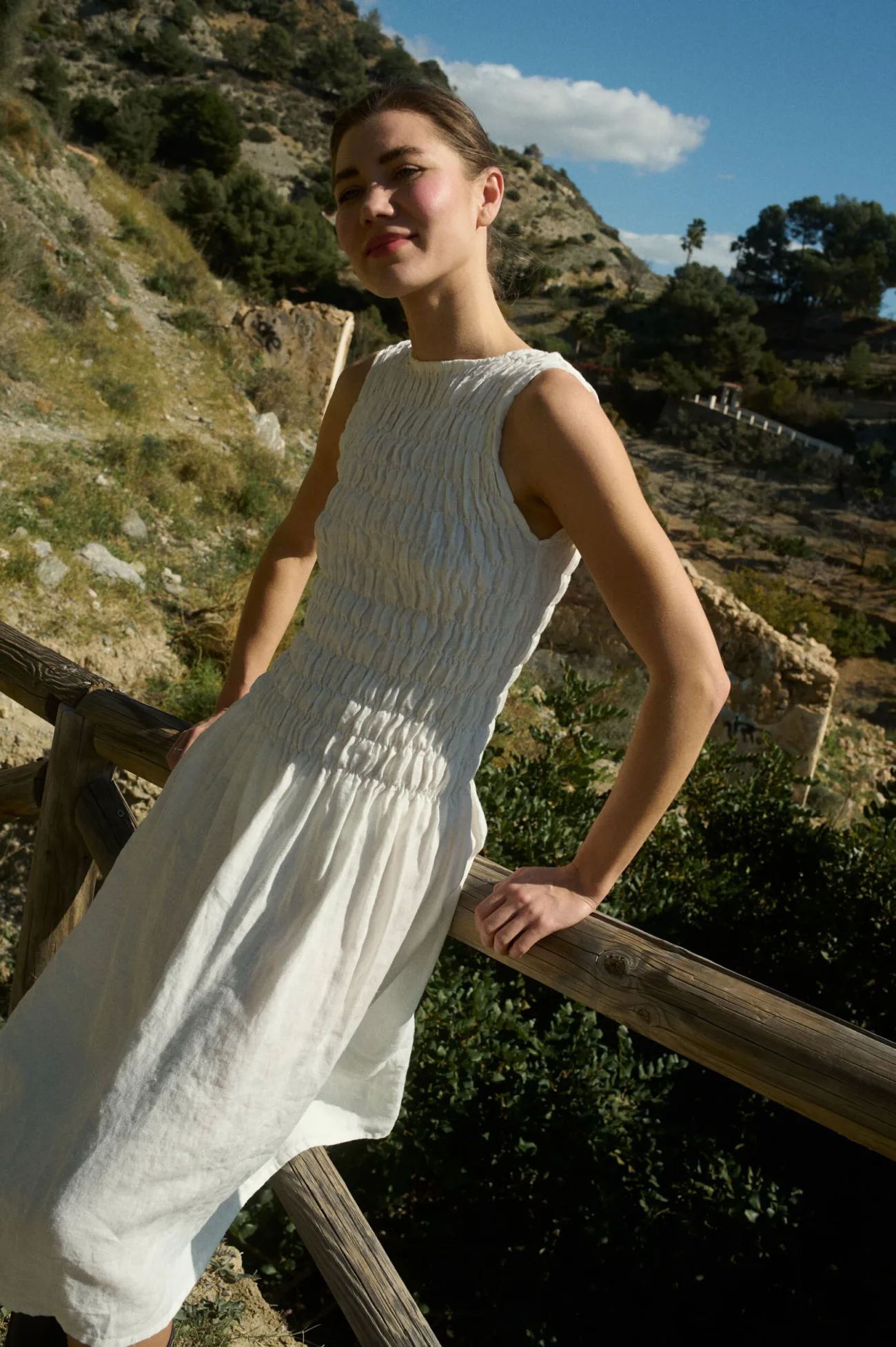 Woman wearing a white linen dress with intricate hand-smocked detailing across the bodice, featuring a sleeveless design, straight neckline, and textured honeycomb-like pattern that extends from the neckline to the waist