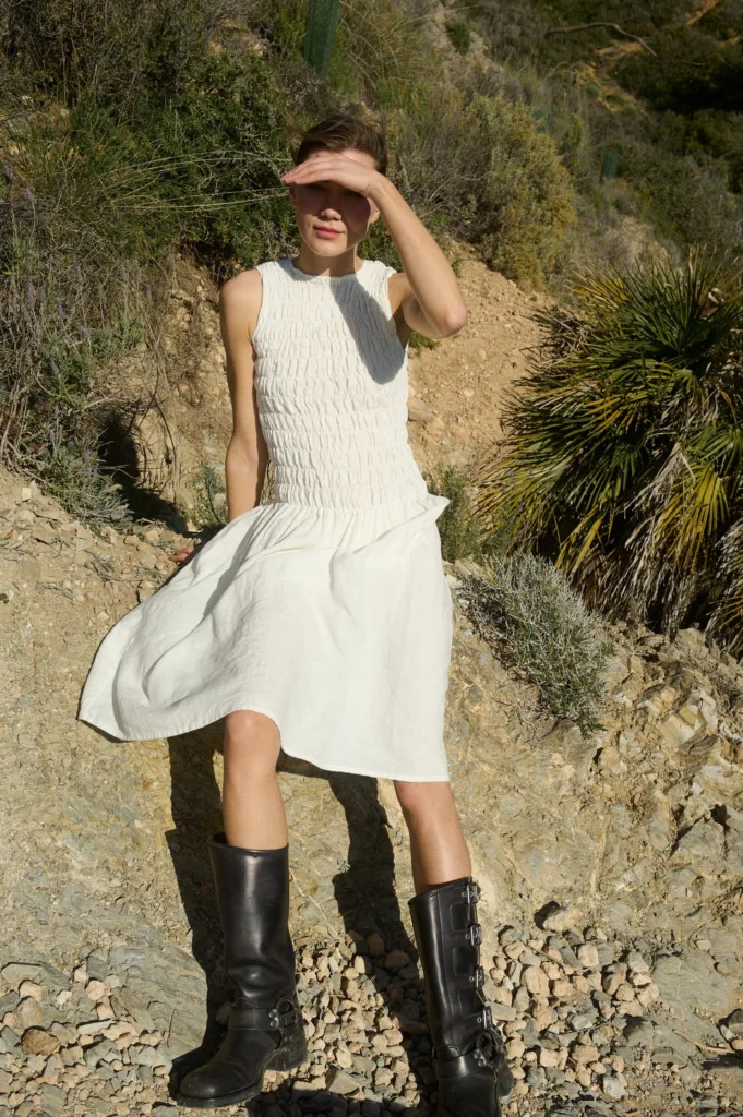 Woman is sitting wearing a knee-length white linen dress with intricate hand-smocked detailing across the bodice, a sleeveless design, straight neckline, and a relaxed A-line skirt that falls just below the knee