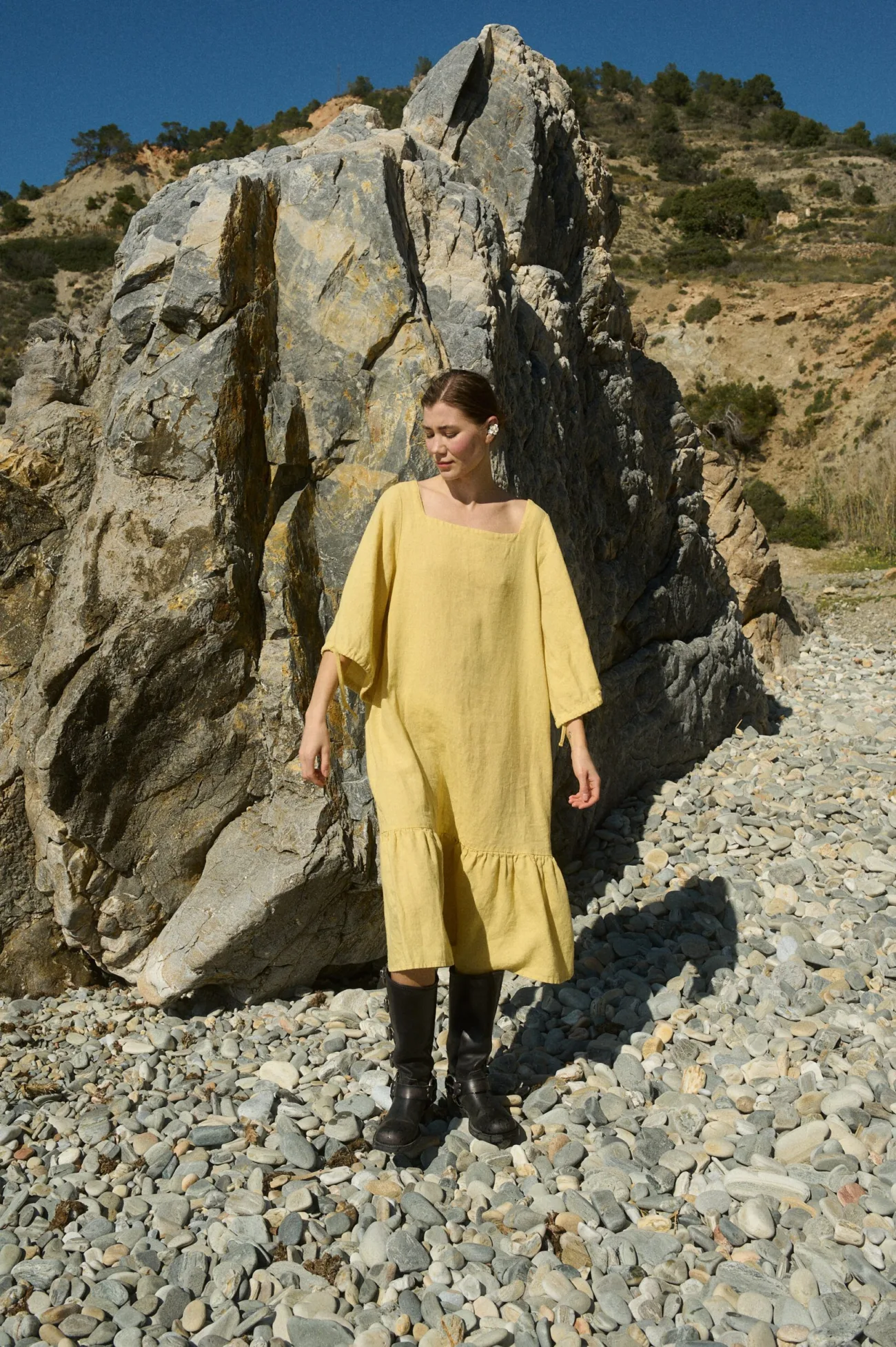 Model showcasing a butter-colored midi linen dress with a relaxed fit, featuring a squared neckline, ¾ sleeves with tie details, and a gently ruffled hem that creates a flowing silhouette