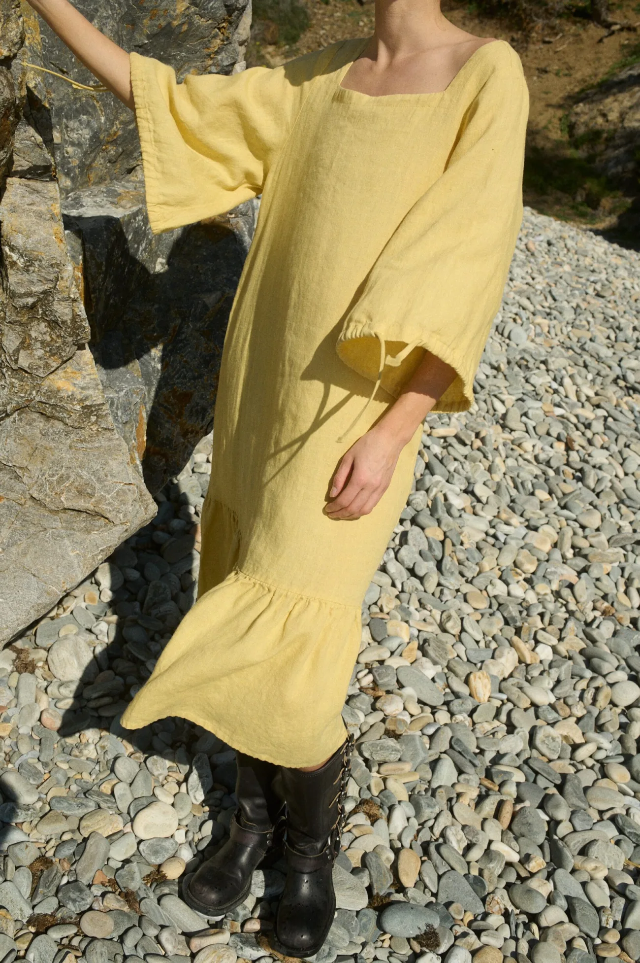 Front view of a butter-colored midi-length oversized linen dress with a wide, squared neckline, ¾ length set-in sleeves, and functional string ties at the cuffs, finished with a softly ruffled skirt hem