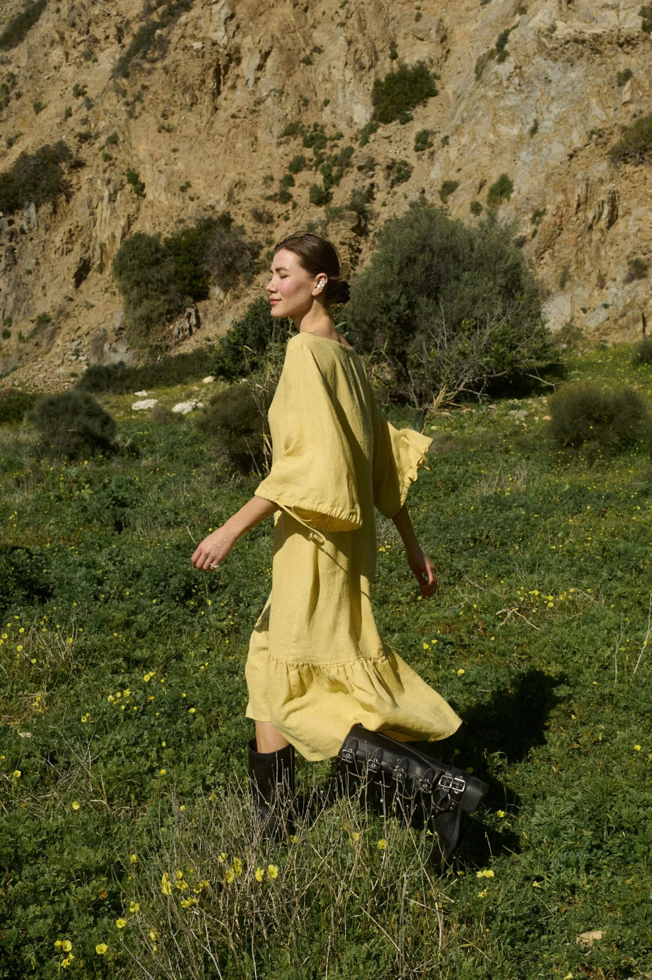 Model walking in a butter-colored midi linen dress with a relaxed fit, featuring a squared neckline, ¾ sleeves with tie details, and a softly ruffled hem that moves with each step