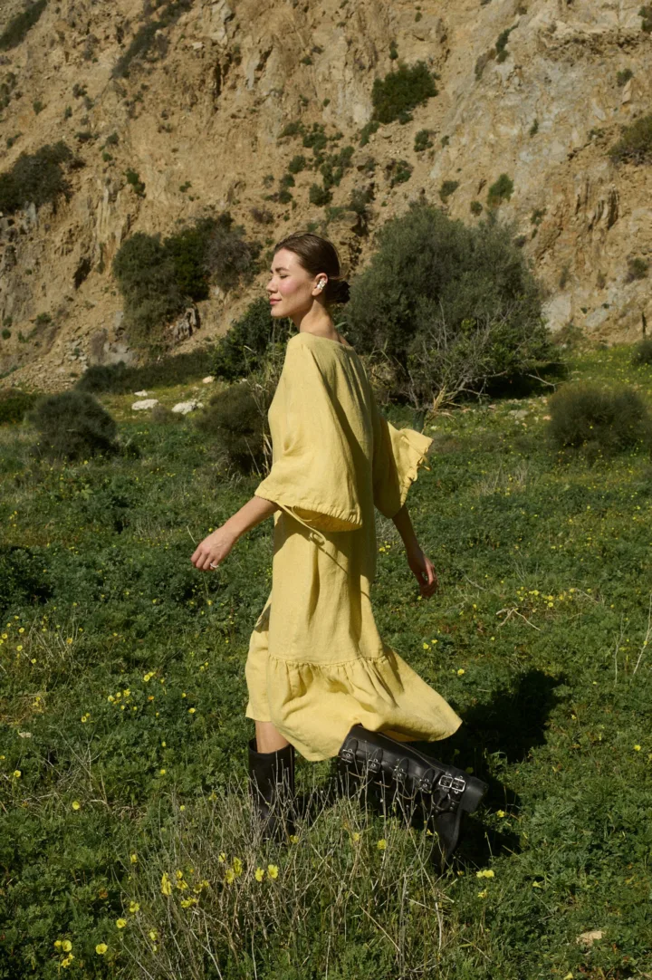 Model walking in a butter-colored midi linen dress with a relaxed fit, featuring a squared neckline, ¾ sleeves with tie details, and a softly ruffled hem that moves with each step