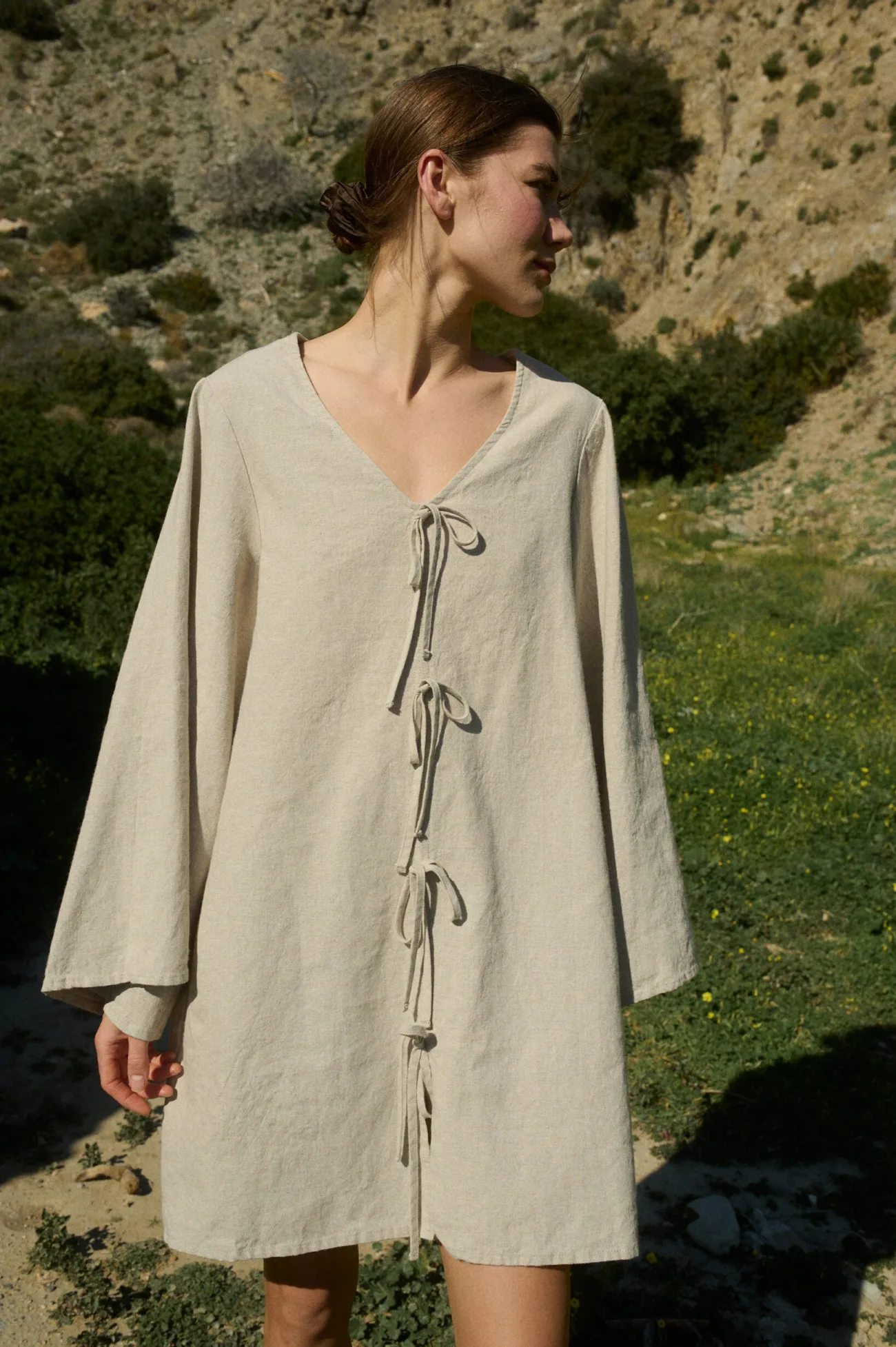 Model showcasing a natural cotton-linen blend mini dress with a deep V-neck, flowing sleeves, wide shoulders, and decorative tie-bows at the front, creating a relaxed A-line silhouette