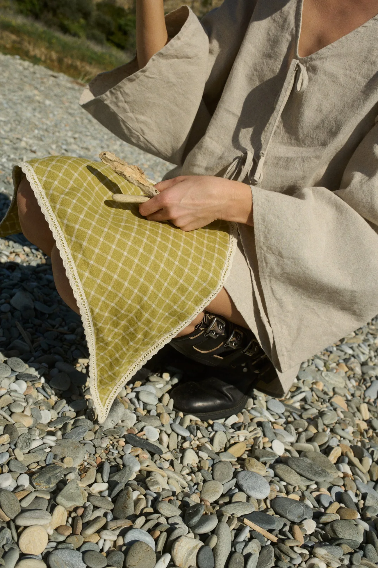 A close-up of a linen bandana in a pickle checks, finished with delicate white lace trim, held by a person sitting on a rocky beach while wearing a natural cotton blend dress