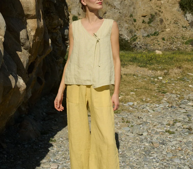 Model in a pale yellow checkered sleeveless linen top featuring a V-neckline, oversized fit, and a single side tie closure, paired with matching butter colored wide-leg pants for a breezy, effortless summer ensemble
