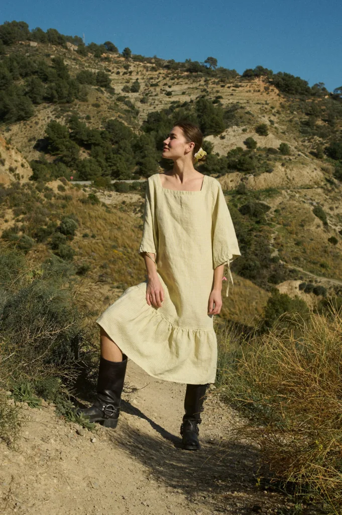 Model wearing a midi light apple checks linen dress with a square neckline, voluminous three-quarter length sleeves gathered at the elbow, and a dramatic drop-waist silhouette with a gathered ruffle at the bottom hem