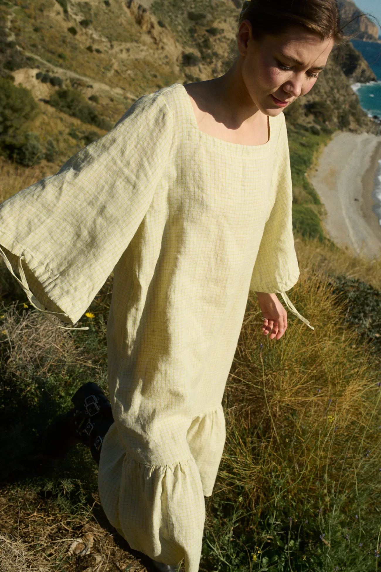 Model wearing an apple checks linen dress with a square neckline, three-quarter length sleeves gathered at the elbows, and a drop-waist silhouette, finished with a ruffled hem