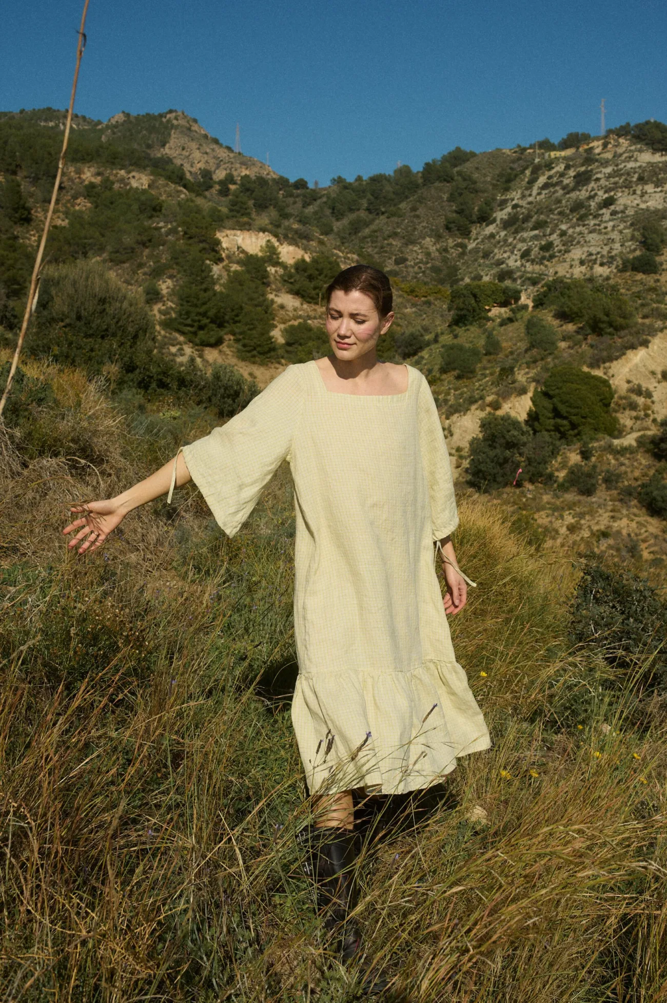 Model wearing an apple checks linen dress featuring a square neckline, voluminous three-quarter length sleeves gathered at the elbow, and a drop-waist silhouette with a gathered ruffle at the hem