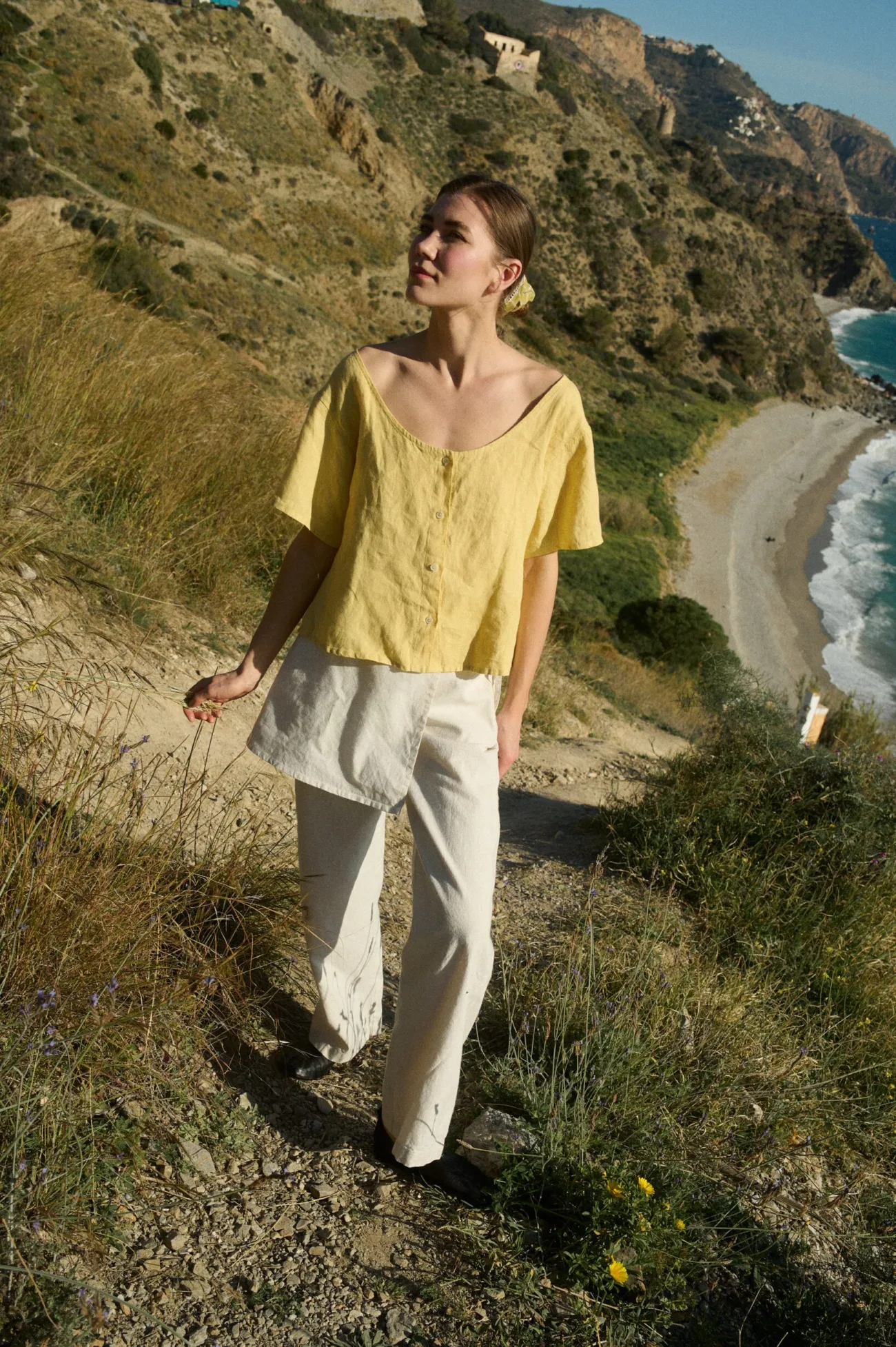 Model standing in a soft, sunlit setting, wearing a butter-yellow linen button-up top with a wide scoop neckline and short sleeves, paired with white wide-leg cotton-blend pants
