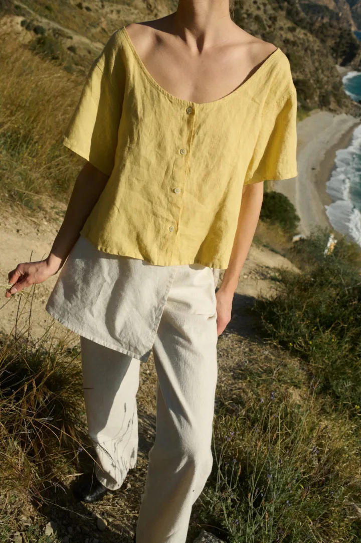 Model wearing a butter yellow linen button-up top with a wide scoop neckline and short sleeves, paired with off white colored wide-leg cotton blend pants