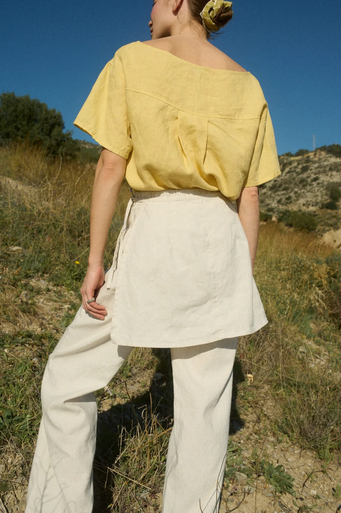 Model seen from the back, wearing a butter-yellow linen button-up top tucked into off white wide-leg cotton-blend pants, which feature an overlapping skirt panel for added dimension