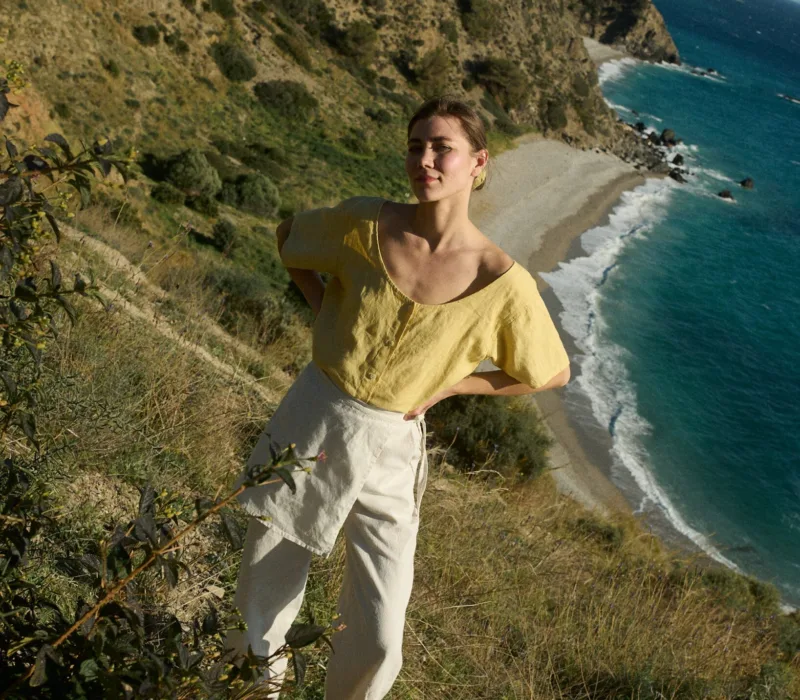 Model wearing a short-sleeved butter-yellow linen button-up top with a wide scoop neckline, tucked into flowing white cotton-blend wide-leg pants with a unique overlapping skirt panel