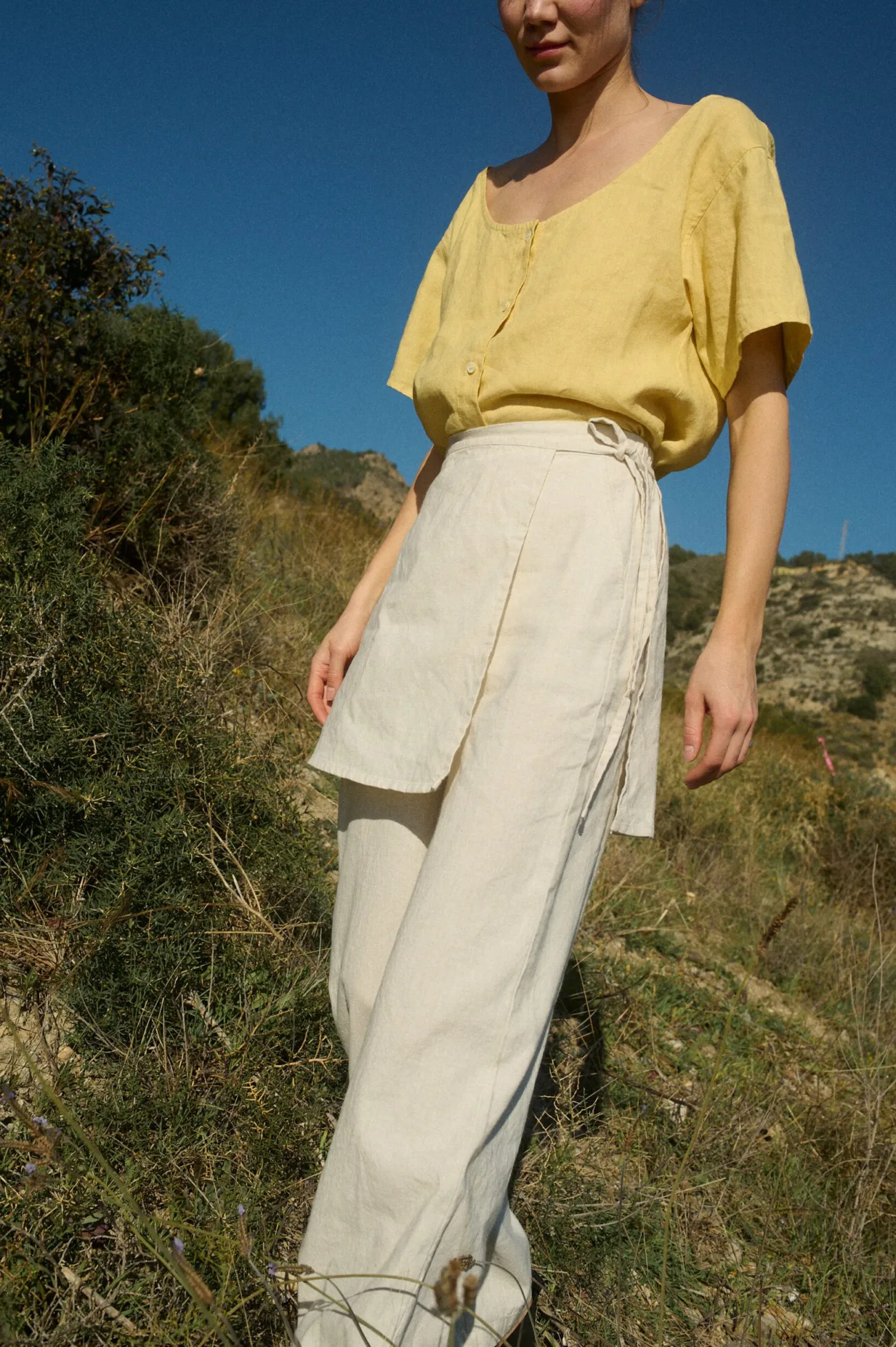 The close-up perspective highlights the delicate details of the sunshine-yellow linen top with its subtle button placket and scoop neckline, paired with airy cream wrap-layer pants that create an effortlessly elegant silhouette