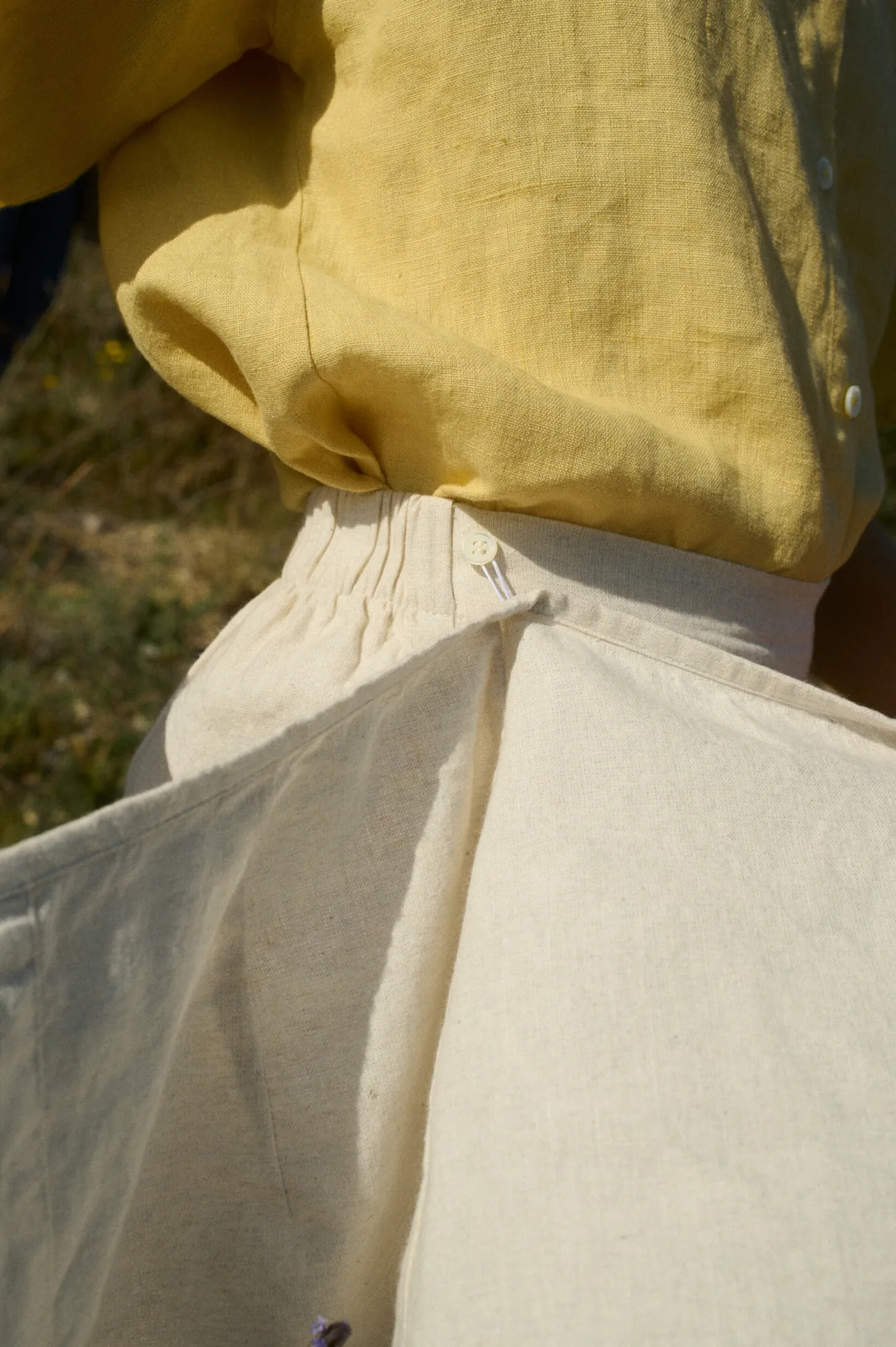 Close-up of a yellow linen top tucked into natural grey cotton-blend bottoms, featuring an elastic waistband and a wrap panel detail