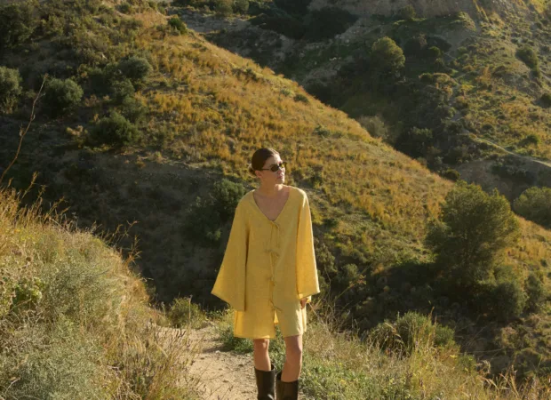 Model wearing a butter yellow linen tunic dress with wide, bell-shaped sleeves, a V-neckline, and a mid-thigh hem, paired with knee-high black leather boots and sunglasses, standing on a hiking trail with golden hills and evergreen trees in the background