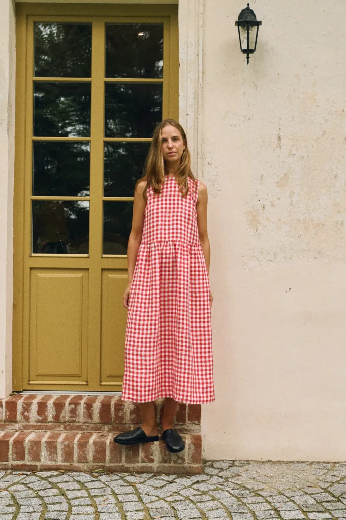 A woman wearing a sleeveless red and white gingham linen dress with a drop waist and gathered midi-length skirt