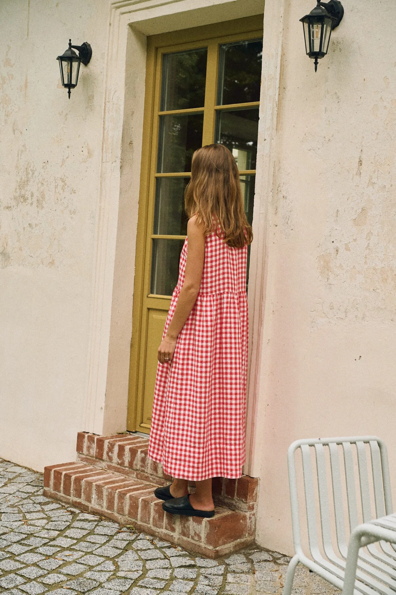 A woman seen from the back, wearing a sleeveless red and white gingham linen dress with a drop waist and a gathered, mid-length skirt.