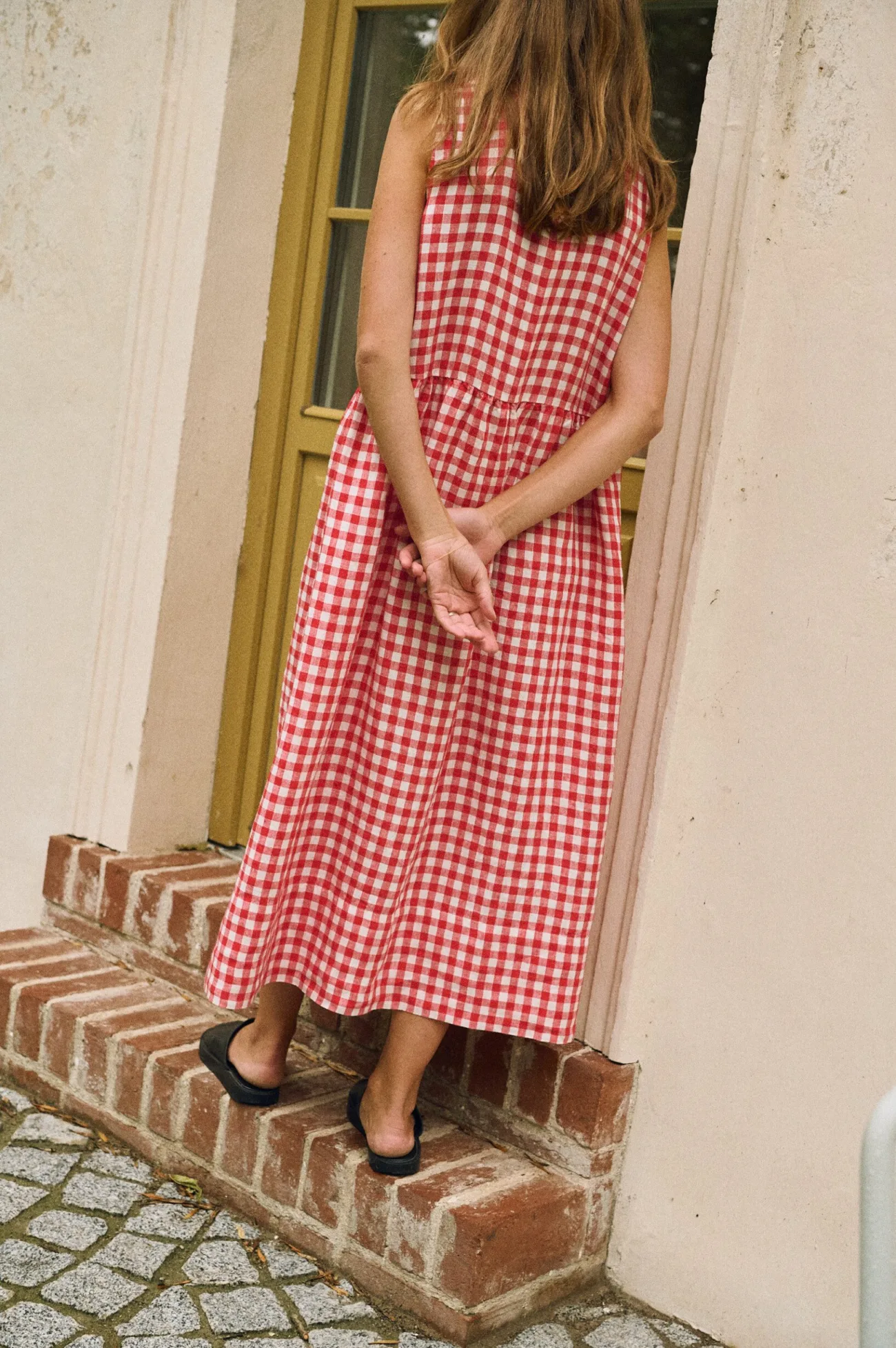 A woman shown from behind, wearing a sleeveless red and white gingham linen dress with a gathered midi-length skirt and a drop waist.