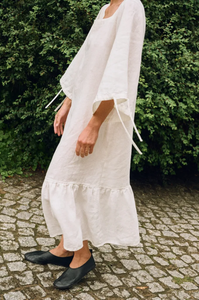 Model wearing an off-white linen midi dress with wide sleeves and ruffle hem, side view highlighting the natural linen drape