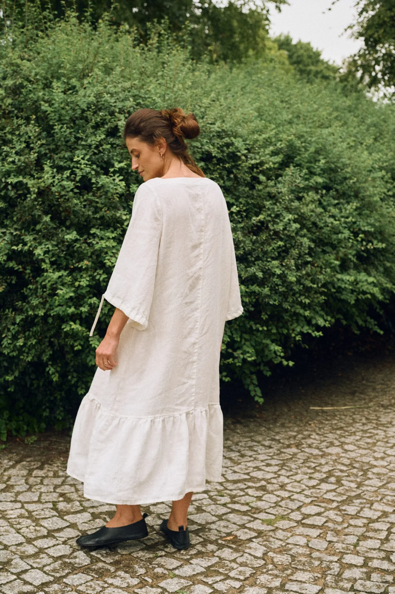 Model wearing an off-white linen midi dress with wide sleeves and ruffle hem, back view highlighting the natural linen drape