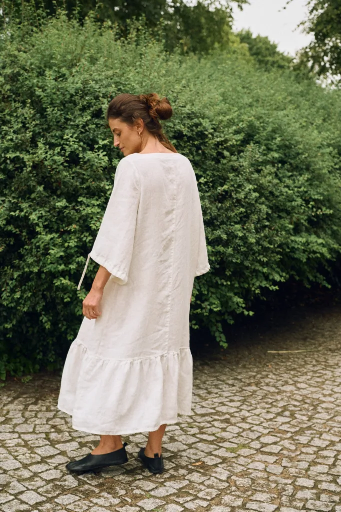 Model wearing an off-white linen midi dress with wide sleeves and ruffle hem, back view highlighting the natural linen drape