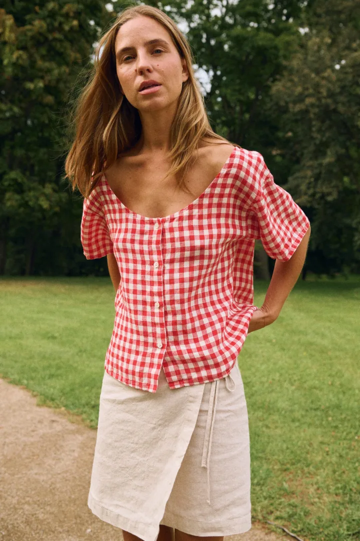 Woman wearing a red gingham linen button-front top with a curved neckline and short sleeves, paired with a natural linen cotton blend wrap skirt with adjustable tie waist