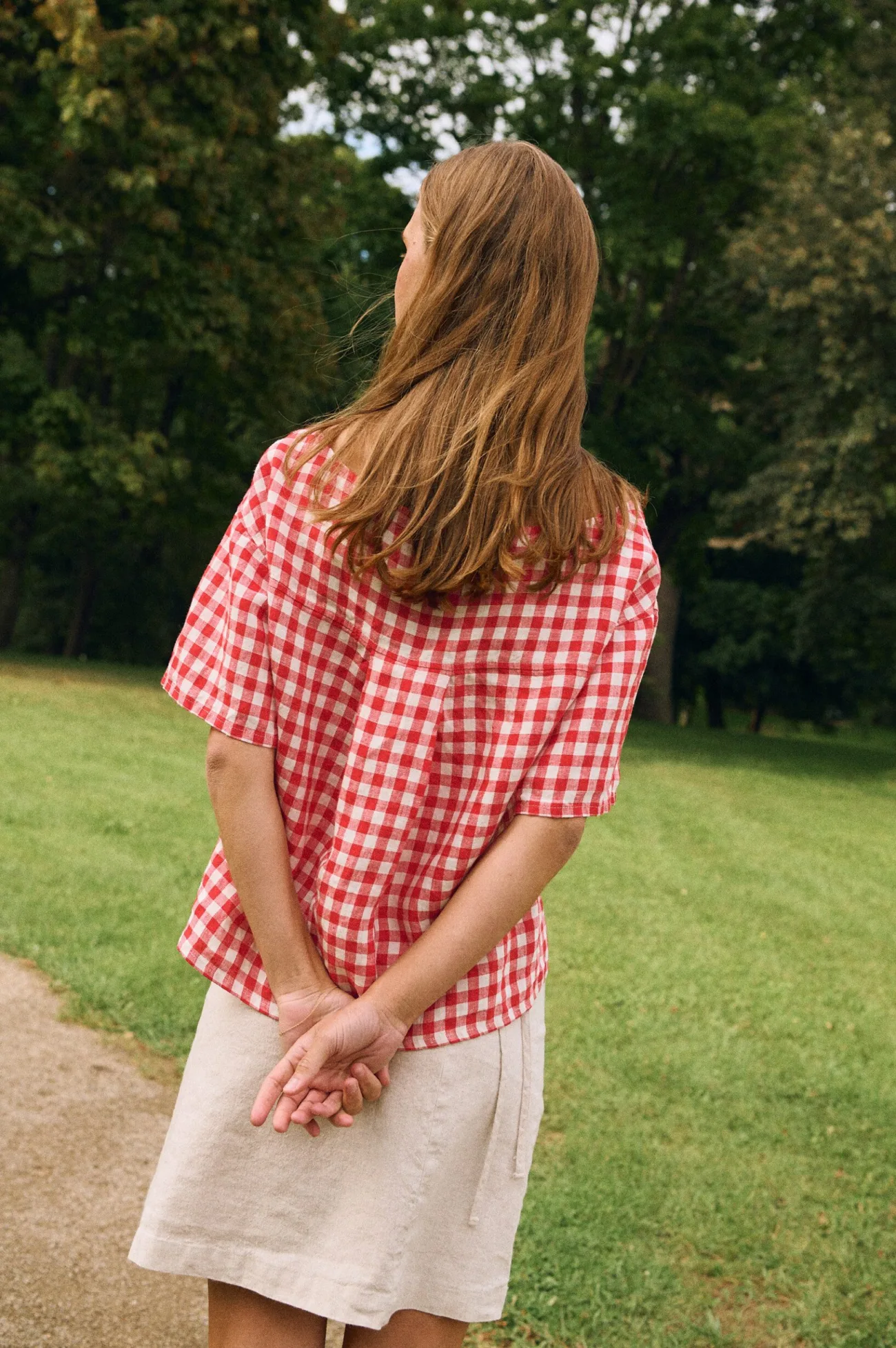 Back view of a model wearing a red gingham linen button-front top with short sleeves, paired with a natural linen cotton blend mini wrap skirt