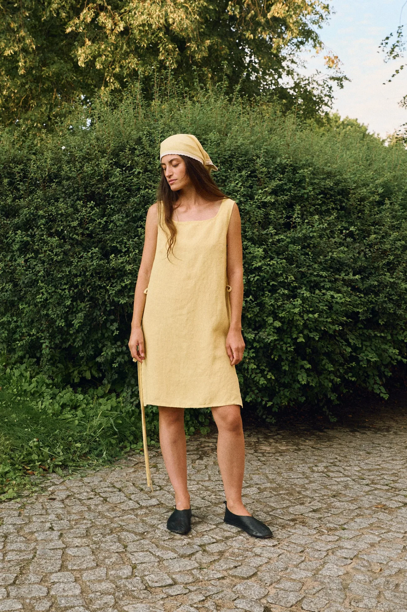 Woman wearing a sleeveless butter yellow linen midi dress with a scoop neckline, side tie accents, and a coordinating linen bandana