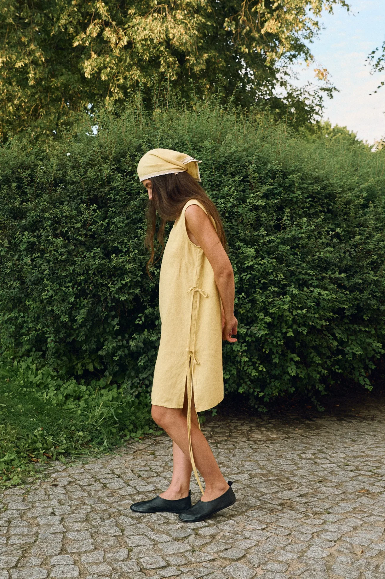 Woman wearing a sleeveless butter yellow linen midi dress with size slits and side tie details, and a matching linen head wrap