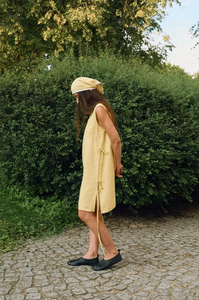 Woman wearing a sleeveless butter yellow linen midi dress with size slits and side tie details, and a matching linen head wrap