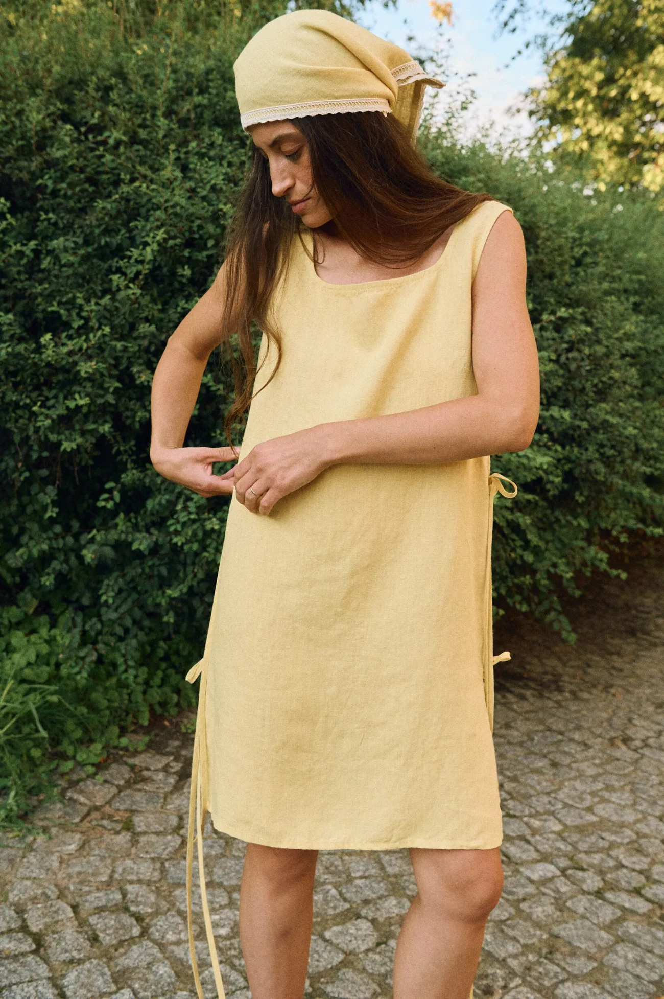 Woman wearing a sleeveless butter yellow linen dress with side ties and a matching head bandana